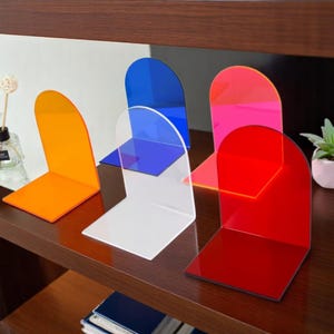 Op de afbeelding: Een verzameling kleurrijke acryl boekensteunen in oranje, blauw, wit, roze en rood. Elke boekensteun heeft een rechthoekige basis en een gebogen bovenkant. Ze zijn geplaatst op een donkere houten plank, met een modern design.