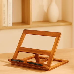 May include: A wooden book stand with a natural finish. The stand is angled with a rectangular base and a frame to hold a book open. The stand has a small ledge with two black rubber grips to hold the book in place. The stand is on a light-colored surface.