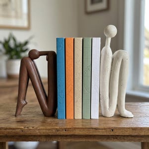 Puede incluir: Un conjunto de cuatro libros con lomos coloridos colocados entre dos sujetalibros con forma humana abstracta. El sujetalibros izquierdo es marrón oscuro y el derecho es blanco roto. Los lomos de los libros son azules, naranjas, beige, verdes y blancos.