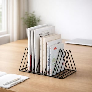 May include: A black metal book organiser holding several books with white covers. The books are arranged in a row, with the spines facing forward. The organiser has a geometric design and sits on a wooden desk.