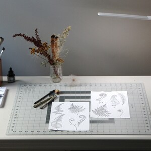 May include: A white desk with a gray cutting mat, a set of watercolor paints, a wooden pencil holder with paintbrushes, a glass jar with dried flowers, a black ink bottle, a white lamp, a black mug, and two sheets of paper with drawings of ferns.