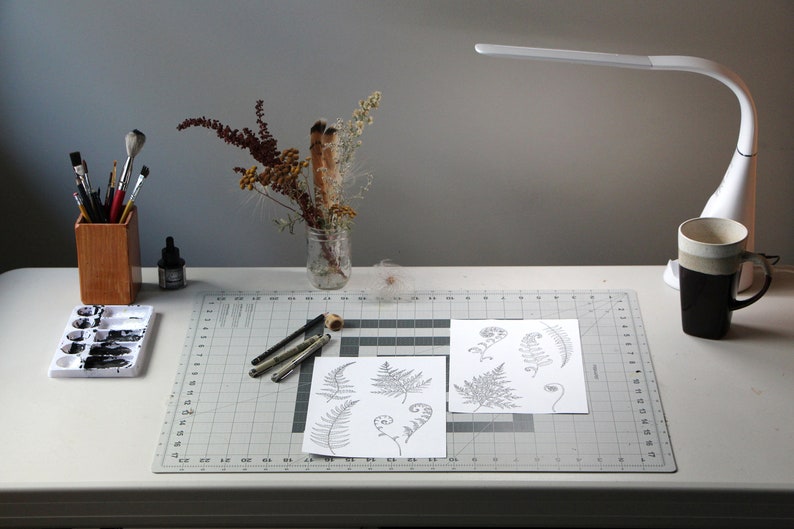 May include: A white desk with a gray cutting mat, a set of watercolor paints, a wooden pencil holder with paintbrushes, a glass jar with dried flowers, a black ink bottle, a white lamp, a black mug, and two sheets of paper with drawings of ferns.