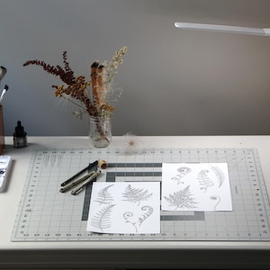 Pu&ograve; includere: Una scrivania bianca con una lampada bianca, un vaso di vetro trasparente con fiori secchi, un portapenne in legno marrone con matite, un tappetino da taglio a griglia bianca con due fogli di carta con disegni a inchiostro nero di felci, una tavolozza di vernice bianca con vernice nera e una tazza bianca con un design bianco e nero.