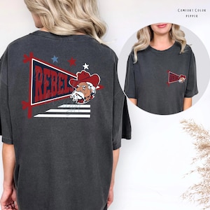 Op de afbeelding: Een antracietgrijs T-shirt met korte mouwen met een grote afbeelding op de achterkant en een kleinere versie op de linkerborst. De afbeelding toont een rode wimpel met het woord REBELS, een cowboy-mascotte en sterren. Tekst luidt COMFORT COLOR PEPPER.
