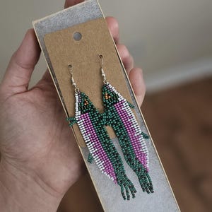 May include: A pair of beaded earrings in the shape of fish, presented in a small box. The earrings feature a design with green, white, and pink beads, with silver-colored ear wires. The fish-shaped earrings are a unique accessory.
