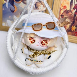Puede incluir: Una muñeca de peluche con cabello claro, ojos en forma de cruz roja y un sombrero de pescador blanco con gafas de sol marrones y una pluma. La muñeca viste un conjunto blanco con una bufanda de leopardo en una silla colgante de macramé.