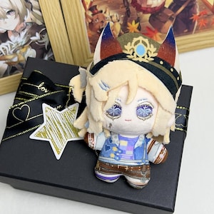Peut inclure: Une poupée en peluche à l'apparence de renard, portant un chapeau et une veste de style militaire. La poupée a des yeux brodés et est posée sur un coffret cadeau noir avec un ruban et une étiquette en forme d'étoile. Des œuvres encadrées sont visibles en arrière-plan.