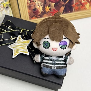 May include: A plush doll with brown hair, green and purple eyes, and a striped shirt. The doll is on a black box with a gold star-shaped tag and a black ribbon with gold text. A framed picture is in the background.