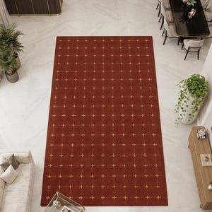 May include: A large rectangular area rug in a deep terracotta red colour featuring a repeating grid pattern of thin, light-coloured lines with small cross symbols at each intersection, centred on a white marble floor in a spacious room.