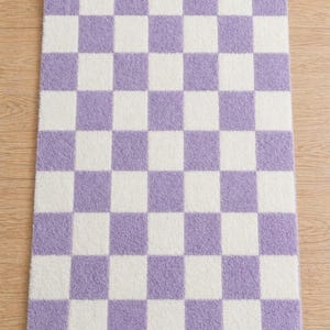 Peut inclure: Un tapis rectangulaire à motif de damier. Le tapis présente des carrés alternés de lavande et de blanc. Le tapis est fait d'une matière douce et moelleuse, et est posé sur un sol en bois.