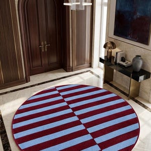 May include: A circular area rug featuring a geometric pattern of alternating horizontal stripes in deep burgundy and light blue. The rug is divided into two distinct sections where the stripes shift direction, creating a split visual effect.
