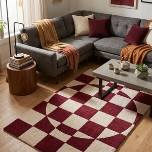 May include: A living room scene featuring a gray sectional sofa with burgundy and cream-colored throw pillows and blankets. A geometric rug with a burgundy and cream checkerboard pattern is on the wooden floor. A side table and a coffee table are also present.