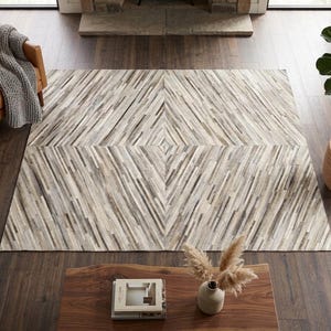 Puede incluir: Una alfombra cuadrada grande con un patrón geométrico de tiras finas multidireccionales en tonos crema, beige, gris y marrón oscuro. Las tiras están dispuestas en un diseño simétrico en forma de diamante que irradia desde el centro.