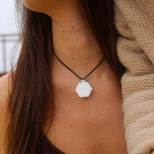 May include: A white hexagonal pendant necklace with a black cord. The pendant has a silver-colored metal accent at the top. The necklace is worn against a person's skin, with a brown top and a beige sweater sleeve in the background.