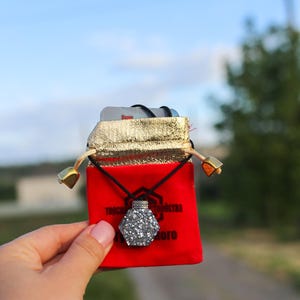 May include: A silver-colored, hexagonal pendant necklace with a sparkling, textured surface hangs from a black cord. The pendant is displayed in a red velvet pouch with gold trim and a drawstring closure. The pouch has text printed on it.