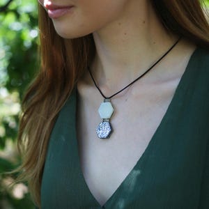 May include: A pendant necklace with two hexagonal pendants. The top pendant is white, and the bottom pendant is a silver and blue glitter design. The necklace is on a black cord. The person is wearing a dark green dress.