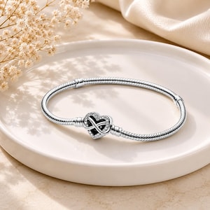 May include: A silver snake chain bracelet featuring a heart-shaped clasp. The clasp is decorated with a central infinity symbol and small clear stones set along the heart's border. The bracelet has a smooth, polished finish and a flexible design.