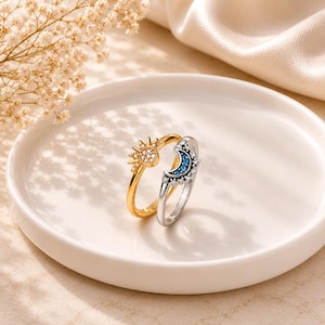 Può includere: Un anello color oro con un motivo a sole con pietre chiare e un anello color argento con un motivo a mezzaluna con pietre blu e chiare. I due anelli sono posizionati insieme, mettendo in risalto i loro distinti design celesti e finiture.