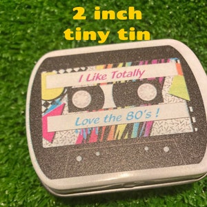 Pu&ograve; includere: Una piccola scatola di latta di 5 cm con un design a cassetta. Il design presenta le scritte "I Like Totally" e "Love the 80's!" in un font retr&ograve;, con motivi geometrici colorati.
