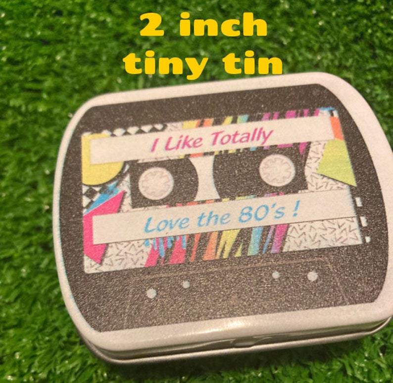 Pu&ograve; includere: Una piccola scatola di latta di 5 cm con un design a cassetta. Il design presenta le scritte "I Like Totally" e "Love the 80's!" in un font retr&ograve;, con motivi geometrici colorati.