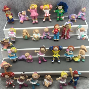 May include: A collection of vintage Cabbage Patch Kids figurines in various poses and outfits. The figurines are brightly colored and feature details like hats, dresses, and accessories. Some are standing, some are crawling, and some are holding items like umbrellas and baseball bats.