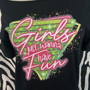 Op de afbeelding: Zwarte off-the-shoulder t-shirt met de tekst "Girls just wanna have Fun" in roze en wit. De tekst staat tegen een neon groen en geel geometrisch ontwerp met kleurrijke spetters. Het shirt wordt getoond op een mannequin.