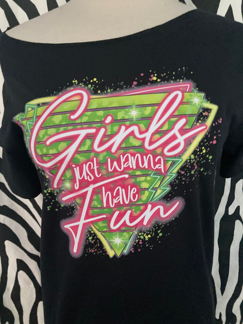 Op de afbeelding: Zwarte off-the-shoulder t-shirt met de tekst "Girls just wanna have Fun" in roze en wit. De tekst staat tegen een neon groen en geel geometrisch ontwerp met kleurrijke spetters. Het shirt wordt getoond op een mannequin.