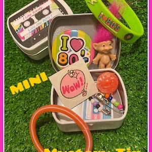 Pu&ograve; includere: Una collezione di oggetti a tema anni '80 in una scatola di latta bianca. Include una scatola a forma di cassetta, un braccialetto verde con la scritta "80s", una bambola troll, un adesivo, un braccialetto arancione e altri piccoli accessori. Il testo "Mystery Tin" &egrave; visibile.