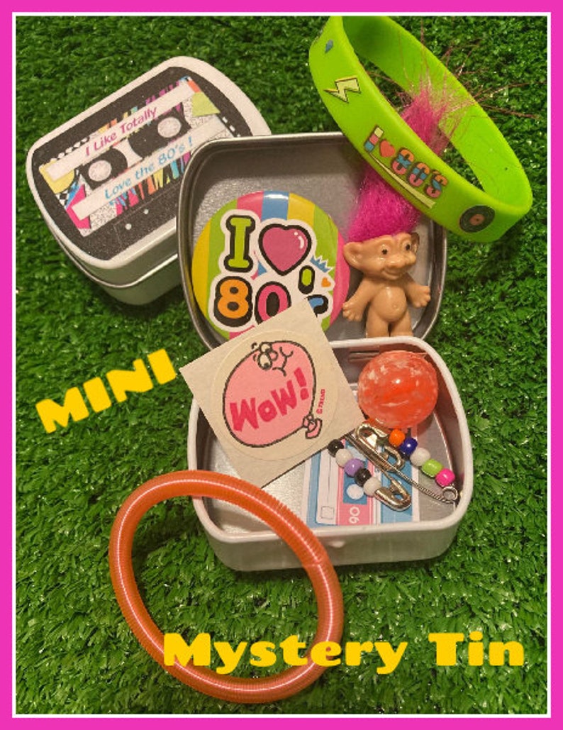 Pu&ograve; includere: Una collezione di oggetti a tema anni '80 in una scatola di latta bianca. Include una scatola a forma di cassetta, un braccialetto verde con la scritta "80s", una bambola troll, un adesivo, un braccialetto arancione e altri piccoli accessori. Il testo "Mystery Tin" &egrave; visibile.