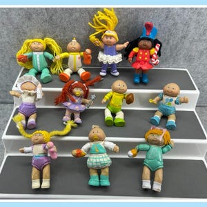 May include: A collection of 10 Cabbage Patch Kids dolls in various outfits and poses. The dolls are made of plastic and have painted faces. They are arranged on a white tiered shelf.