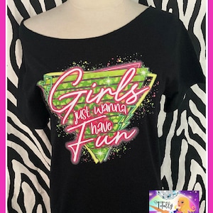 Op de afbeelding: Zwarte off-the-shoulder t-shirt met de tekst "Girls just wanna have fun" in roze en groen. De afbeelding is in een geometrisch ontwerp. Het shirt wordt gepresenteerd op een mannequin met een zebraprint achtergrond.