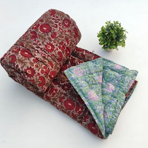 May include: A folded cotton quilt featuring a deep red and brown floral block print on one side and a contrasting light green and blue floral pattern on the reverse. The textile has visible quilted stitching throughout the fabric surface.
