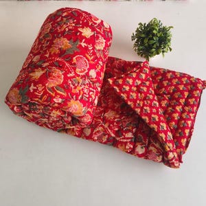 Op de afbeelding: Een gevouwen rode quilt met een herhalend bloemenpatroon in tinten oranje, geel en grijs. De stof heeft een doorgestikte textuur met zichtbare stiksels. Een kleine, ronde, groene kunstplant staat naast het gevouwen textiel.