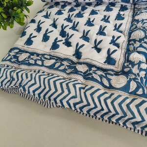 Könnte beinhalten: Eine weiße Steppdecke mit einem sich wiederholenden Muster aus dunkelblauen Kaninchen. Der Rand zeigt ein florales Motiv in Blau und Weiß, ergänzt durch einen Abschnitt mit blau-weißen Zickzackmustern und einem schmalen blau-weiß gestreiften Rand.
