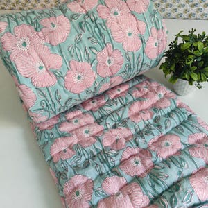 May include: A quilted blanket featuring a repeating pattern of light pink flowers with dark pink line details and green stems on a light teal background. The fabric has a soft, stitched texture throughout, showcasing a floral botanical design.