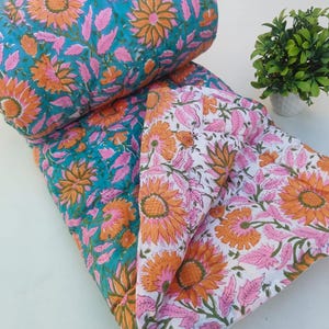 Könnte beinhalten: Eine gesteppte Decke mit einem Blumenmuster aus orangefarbenen Sonnenblumen und rosa Blättern. Eine Seite hat einen leuchtend türkisfarbenen Hintergrund, während die andere Seite einen weißen Hintergrund aufweist. Der Stoff ist durchgehend gesteppt.
