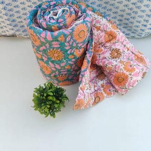 May include: A rolled cotton quilt featuring a vibrant floral pattern with orange sunflowers and pink leaves on a teal background, with a reverse side showing the same floral design in orange and pink on a white background.