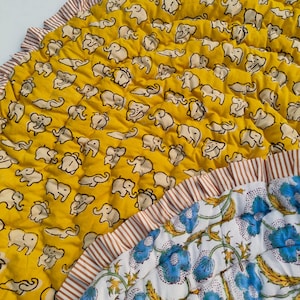 May include: A quilted circular mat featuring a mustard yellow section with a repeating pattern of small white elephants and a white section with blue floral motifs. The edges are finished with a brown and white striped ruffled fabric trim.