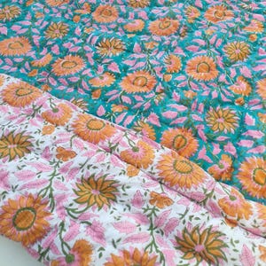 May include: A quilted blanket featuring a repeating floral pattern of orange sunflowers and pink leaves. One side has a vibrant teal background, while the folded corner reveals a white background with the same floral print and visible stitching.