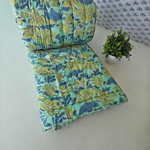 Puede incluir: Una colcha plegada con un fondo verde menta claro y un patrón repetitivo de flores grandes amarillas y hojas azul oscuro. La tela presenta costuras lineales visibles en toda su extensión, creando una textura acolchada en la superficie.