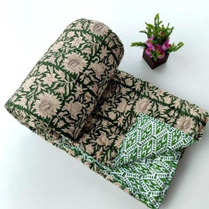 Könnte beinhalten: Eine gefaltete Steppdecke mit dunkelgrünem Hintergrund und beigem Blumenmuster auf der einen Seite sowie einem weiß-grünen geometrischen Rautenmuster auf der Rückseite. Der Stoff weist durchgehend sichtbare Steppnähte auf.