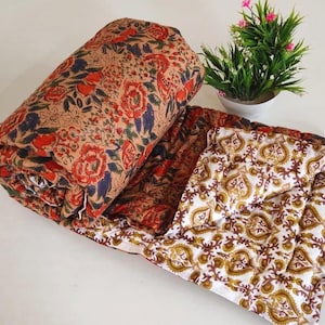 Puede incluir: Una manta acolchada con fondo color canela y un denso estampado floral en rojo, naranja y azul oscuro. El reverso de la manta doblada muestra una tela blanca con un estampado botánico repetitivo en tonos marrón y dorado.