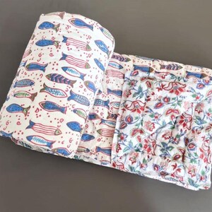 Può includere: Una coperta trapuntata con sfondo bianco e un motivo ripetuto di pesci blu, rossi e marroni su un lato, e un motivo floreale a contrasto con disegni botanici rossi, blu e verdi sul retro, ripiegata per mostrare entrambe le stampe.