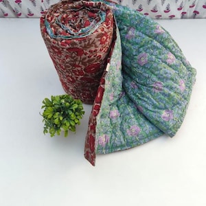 May include: A rolled quilted blanket featuring a deep red and brown floral block print on the exterior and a light green fabric with a purple and blue floral pattern on the interior, showcasing visible stitching throughout the textile surface.