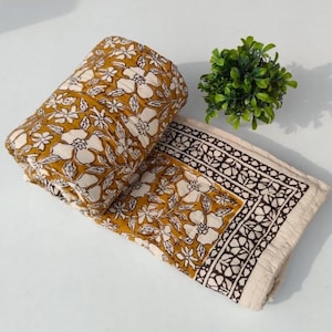 May include: A folded mustard yellow quilt featuring a white floral pattern and a dark brown geometric border. The fabric has visible stitching throughout, creating a quilted texture. A small green potted plant is positioned near the quilt.
