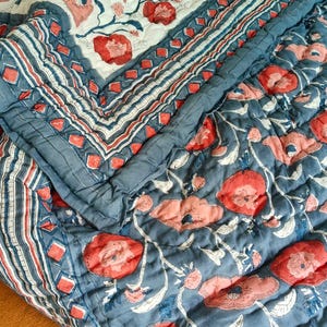 May include: A quilted cotton bedspread featuring a floral pattern with red and pink poppies on a blue background, contrasted by a section with a white background. The edges are finished with a decorative border of blue and red geometric shapes.