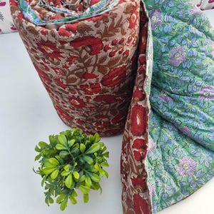 Op de afbeelding: Een opgerolde gewatteerde deken met een bloemenpatroon met rode en bruine bloemen op een beige achtergrond aan de ene kant, en een groene achtergrond met blauwe en paarse bloemen aan de andere kant, met gedetailleerde stiksels in de stof.