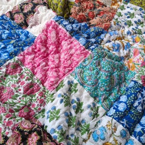 May include: A quilted blanket featuring a patchwork of square fabric panels in various colors including pink, blue, green, black, and yellow. Each square displays a distinct floral or paisley print pattern with a textured, puckered surface finish.