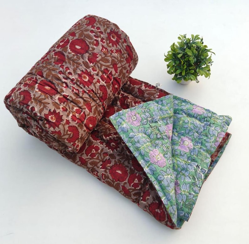 May include: A folded cotton quilt featuring a deep red and brown floral block print on one side and a contrasting light green and blue floral pattern on the reverse. The textile has visible quilted stitching throughout the fabric surface.