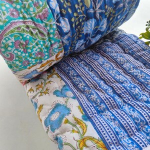 May include: A quilted cushion featuring a patchwork design with various floral and paisley patterns in shades of blue, white, pink, and green. The fabric is stitched with a grid pattern, showcasing multiple distinct textile sections joined together.
